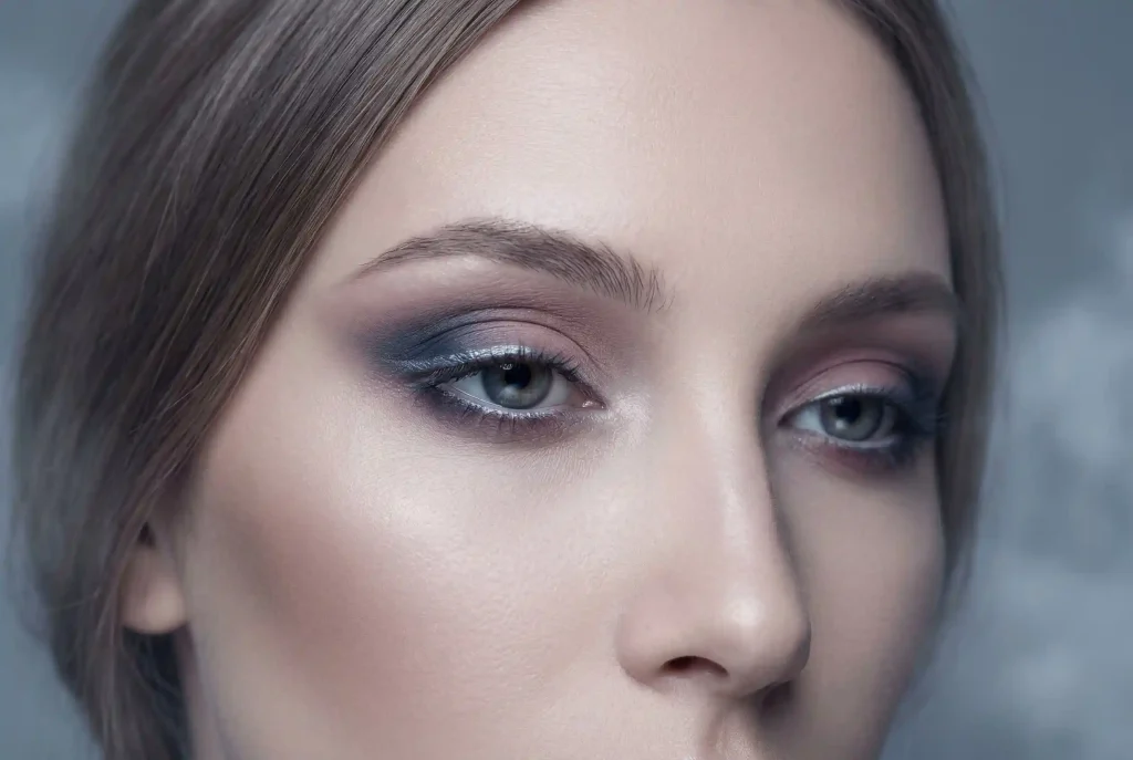 Jak dobrać cienie do powiek? Zobacz, jak dobrać cienie pod kolor oczu i jak pracować cieniami, by powieka zachwycała! Zbliżenie twarzy modelki z makijażem oczu w chłodnych, zgaszonych tonach dymnego granatu i taupe, idealnie dobranym dla letniego typu urody i jego mglistej elegancji.