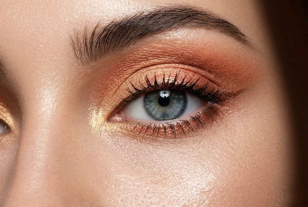 Jakie cienie do powiek do szarych oczu wybrać? Sprawdzony poradnik osobistej stylistki Zbliżenie na szare oko modelki, które pod wpływem nałożonych na powiekę kontrastowych miedzianych, brzoskwiniowych i złotych cieni, zyskuje intensywny, chłodny błękitny odcień.