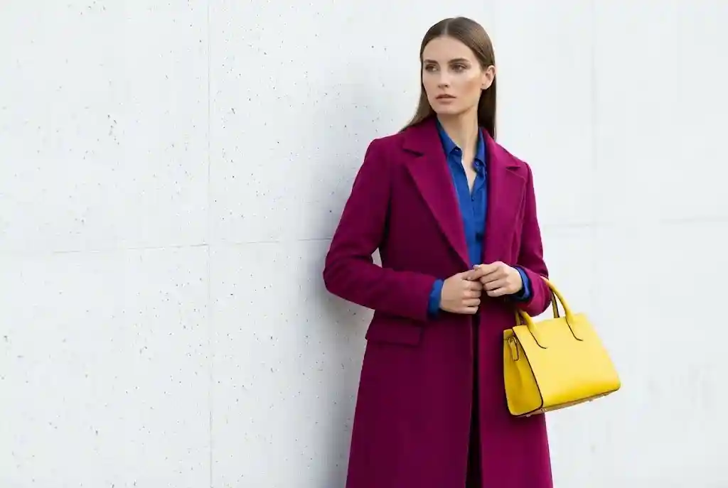 Color blocking — jak łączyć kolory kontrastowe w stylizacji? Modelka prezentująca na sobie regułę 60-30-10 w color blockingu, mająca na sobie dominujący w 60% magentowy płaszcz jako bazę, wspierający musztardowy golf oraz wyrazistą cyjanową torebkę stanowiącą 10% akcentu.